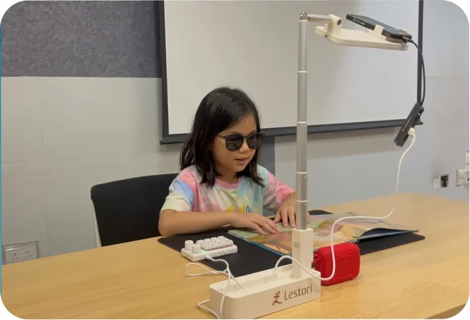 A child in dark glasses reads a picture book by touch, with a braille keyboard, a speaker, and a phone on a Lestori stand